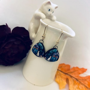 May include: A pair of dangle earrings featuring a whimsical design. Each earring showcases a dark blue, triangular shape with a house, trees, and a white cat illustration. The earrings have gold-colored hooks and small beaded accents.