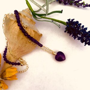 May include: A gold chain necklace with a purple heart pendant and a strand of purple beads. The necklace is lying on a seashell with lavender flowers in the background.