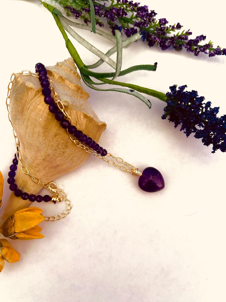 May include: A gold chain necklace with a purple heart pendant and a strand of purple beads. The necklace is lying on a seashell with lavender flowers in the background.