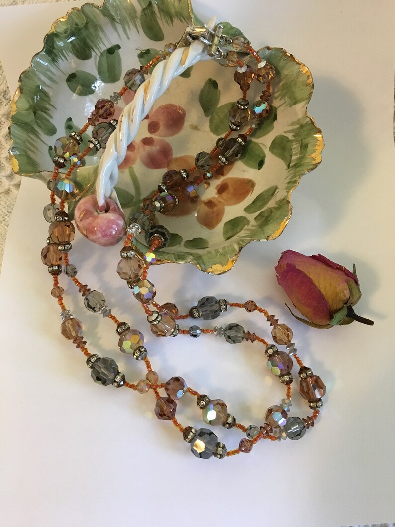 May include: A multi-strand necklace with brown, gray, and clear iridescent beads. The necklace features a pink, round bead in the center.