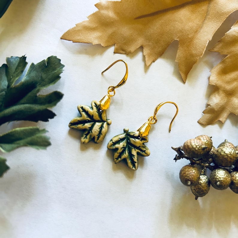 May include: A pair of gold-colored leaf earrings with green and cream-colored details. The earrings are displayed on a white surface with decorative leaves and acorns in the background. The earrings have a hook closure.
