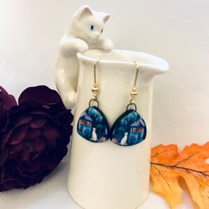May include: A pair of whimsical earrings featuring a house and cat design. The earrings have a blue and white color scheme with gold-colored ear wires. The earrings are displayed on a white ceramic pitcher with a white cat figurine.