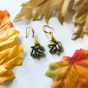 May include: A pair of gold-colored earrings with leaf-shaped pendants. The leaves are green with gold accents, and the earrings have a hook closure. The background features colorful autumn leaves in shades of orange, yellow, and brown.