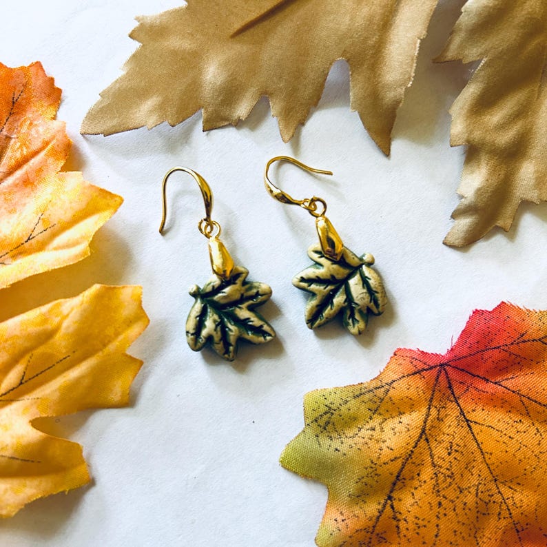 May include: A pair of gold-colored earrings with leaf-shaped pendants. The leaves are green with gold accents, and the earrings have a hook closure. The background features colorful autumn leaves in shades of orange, yellow, and brown.