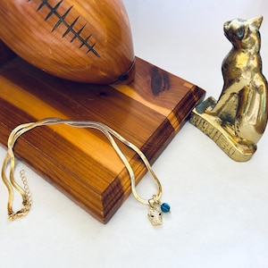 May include: A gold-colored necklace with a pendant shaped like a panther's head, adorned with small black stones and a blue bead. The necklace rests on a wooden surface, alongside a wooden football and a brass cat statue.
