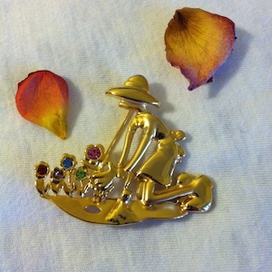 May include: Gold-toned brooch depicting a person gardening. The person is wearing a hat and holding a gardening tool. The brooch features colorful rhinestones representing flowers.