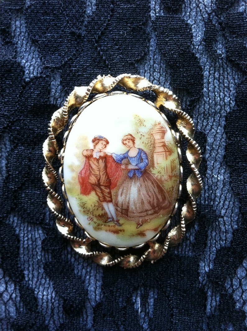 K&ouml;nnte beinhalten: Eine goldfarbene Brosche mit einem wei&szlig;en ovalen Kamee, der eine gemalte Szene von zwei Personen in einem Garten zeigt. Der Kamee ist von einem gedrehten Goldrahmen umgeben. Der Text "Fragonard" ist am unteren Rand des Kamees sichtbar.