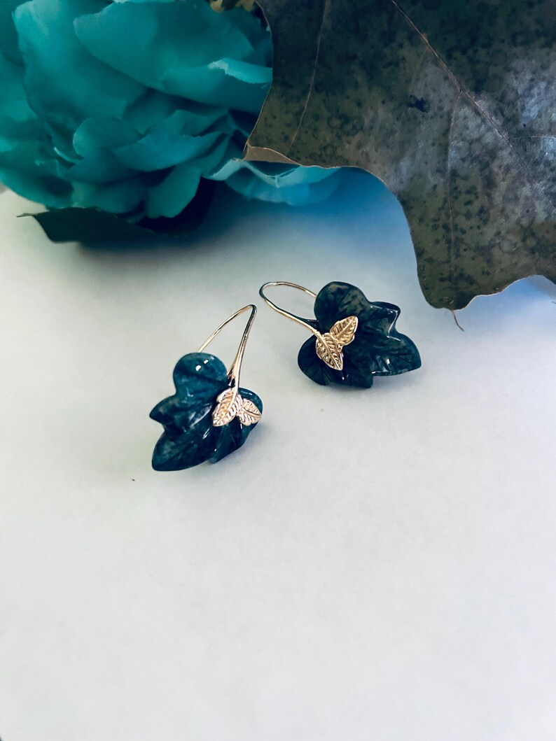 May include: A pair of dark green leaf-shaped earrings with gold accents. The earrings are made of a textured material and have a delicate, feminine design.