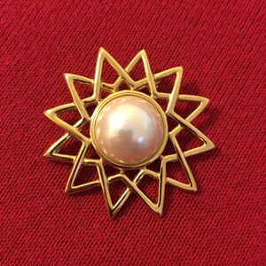 May include: A gold-toned brooch with a starburst design. The brooch features a large, round, pearlescent center stone. The starburst design has pointed edges. The brooch is set against a red background.