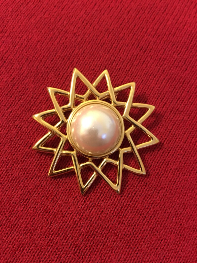 May include: A gold-toned brooch with a starburst design. The brooch features a large, round, pearlescent center stone. The starburst design has pointed edges. The brooch is set against a red background.