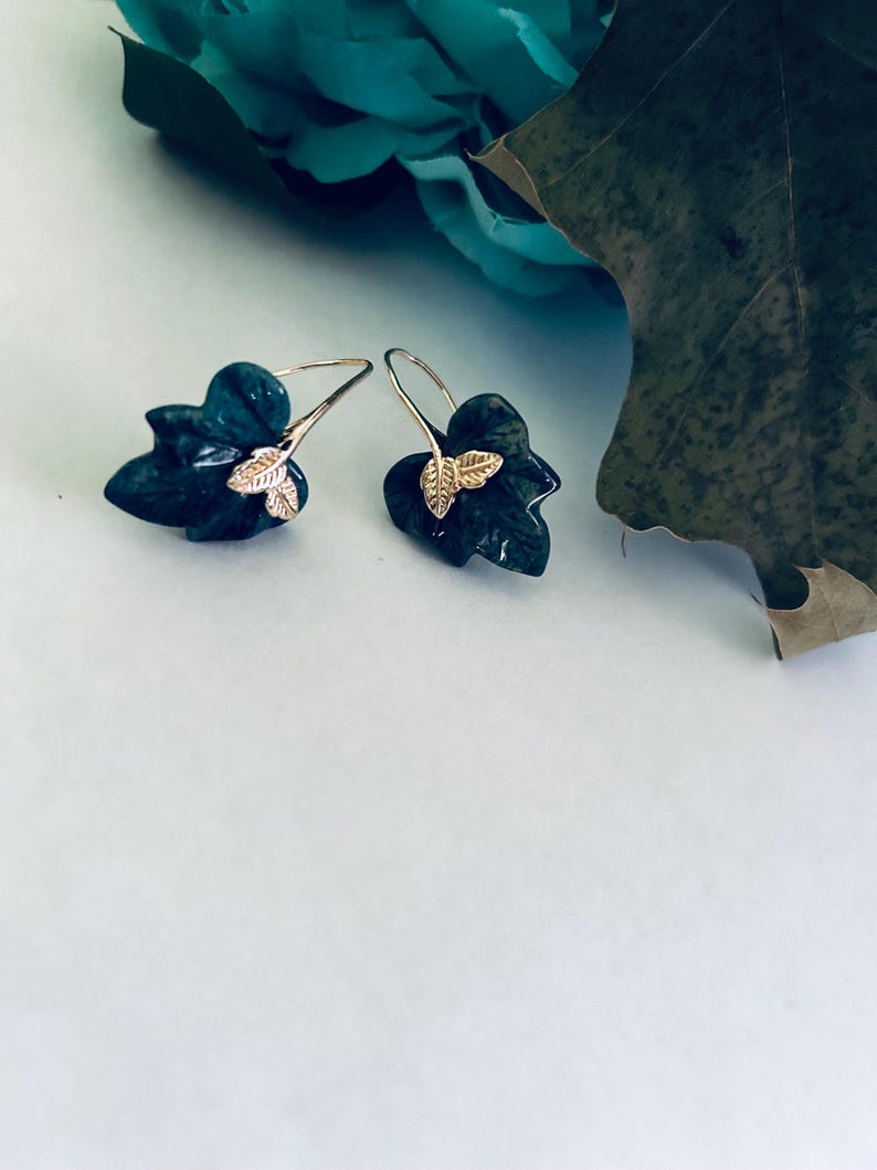 May include: A pair of silver earrings with a green leaf design and gold accents. The leaves are detailed and have a realistic look.
