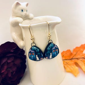May include: A pair of dangle earrings with a whimsical design. Each earring features a dark blue background with a white cat, a house, and trees. The earrings are suspended from gold-colored hooks.
