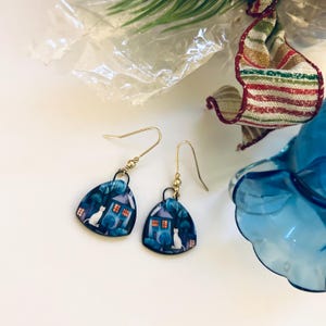 May include: A pair of dangle earrings with a whimsical design. Each earring features a blue, house-shaped pendant with a white cat, a house with lit windows, and trees. The earrings have gold-colored ear wires.