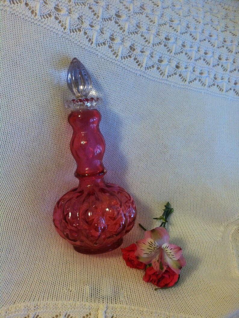 May include: A pink glass bottle with a clear glass stopper. The bottle has a textured surface and is shaped like a gourd. The bottle is sitting on a white crocheted surface with a pink flower.