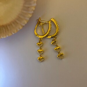 May include: Gold hoop earrings with a dangling chain of four heart-shaped charms. Each heart is accented with small, clear stones.