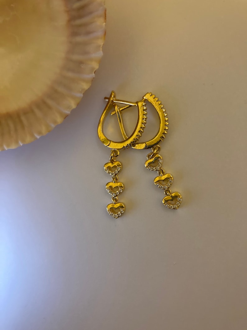 May include: Gold hoop earrings with a dangling chain of four heart-shaped charms. Each heart is accented with small, clear stones.