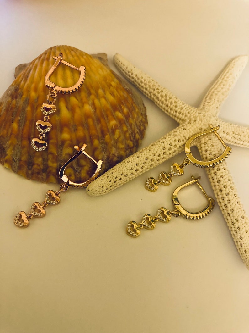 May include: A pair of gold hoop earrings with dangling charms. Each charm is a heart-shaped pendant with clear sparkling stones.