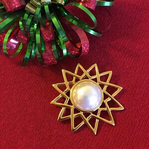 May include: A gold-colored starburst brooch with a large, round, pearlescent center. The brooch is set against a red background, with a decorative bow of green, red, and gold ribbons in the upper left corner. A festive accessory.