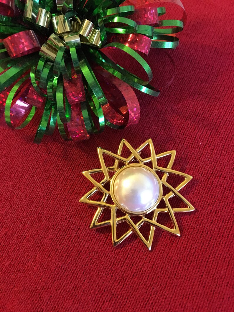 May include: A gold-colored starburst brooch with a large, round, pearlescent center. The brooch is set against a red background, with a decorative bow of green, red, and gold ribbons in the upper left corner. A festive accessory.
