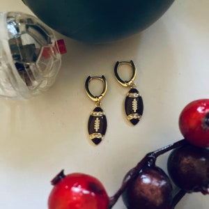May include: A pair of gold hoop earrings with a brown football charm. The football charm has white stitching and small clear rhinestones.
