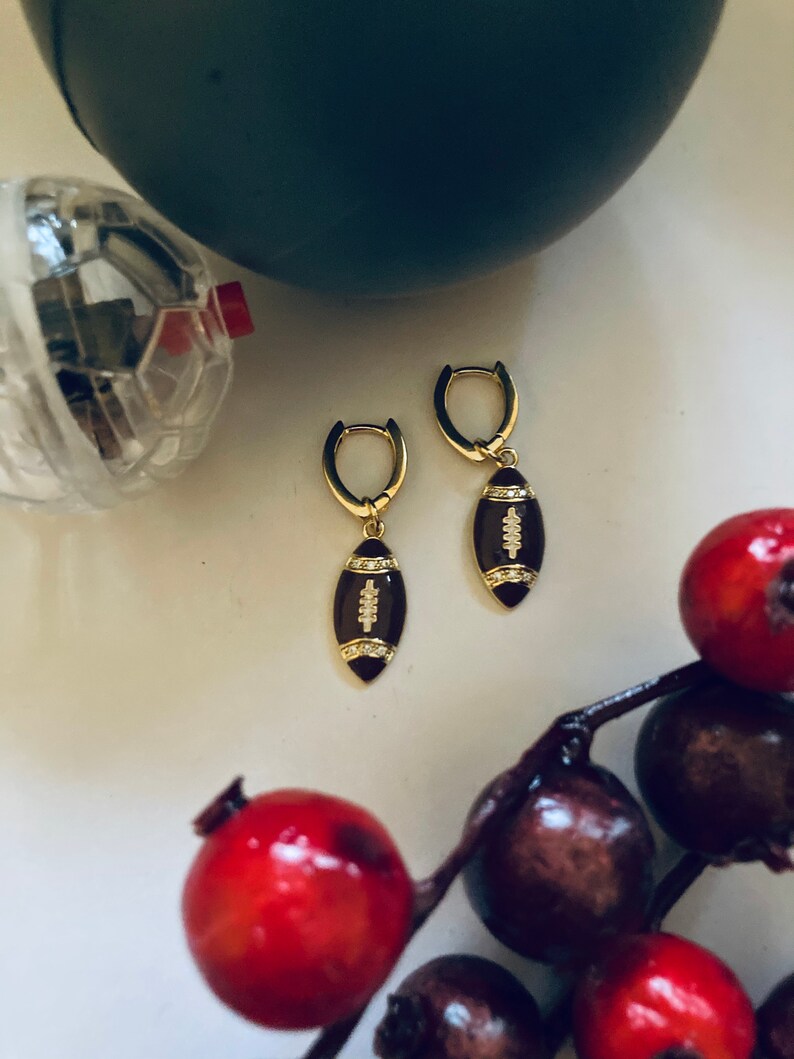 May include: A pair of gold hoop earrings with a brown football charm. The football charm has white stitching and small clear rhinestones.