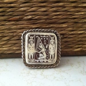 May include: A silver-toned square brooch with a white cameo featuring an Egyptian-inspired design. The cameo depicts a woman playing a harp with two figures standing on either side. The brooch has a rope-like border.