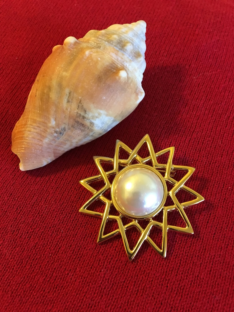 May include: A gold-toned starburst brooch with a large, round, pearlescent center stone. A natural seashell with a light brown and cream color is next to the brooch. The background is a solid red color.