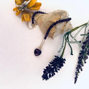 May include: A delicate gold chain necklace with a small heart-shaped amethyst pendant and black beads. The necklace is displayed on a seashell with dried flowers.