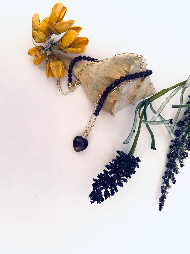 May include: A delicate gold chain necklace with a small heart-shaped amethyst pendant and black beads. The necklace is displayed on a seashell with dried flowers.