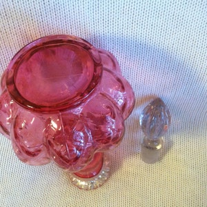 May include: A vintage, ruby-colored glass perfume bottle with a flower-shaped body and a clear, faceted stopper. The bottle has a clear, decorative base. The stopper is a clear, teardrop shape. The bottle is likely from the mid-20th century.