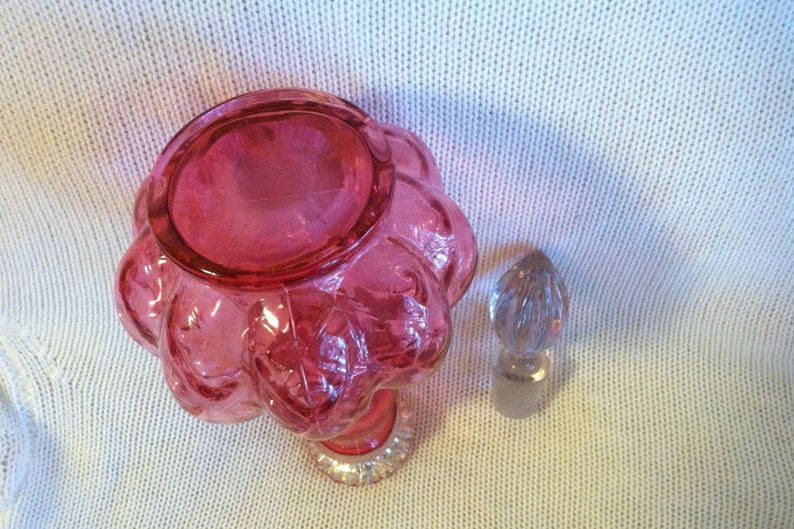May include: A vintage, ruby-colored glass perfume bottle with a flower-shaped body and a clear, faceted stopper. The bottle has a clear, decorative base. The stopper is a clear, teardrop shape. The bottle is likely from the mid-20th century.