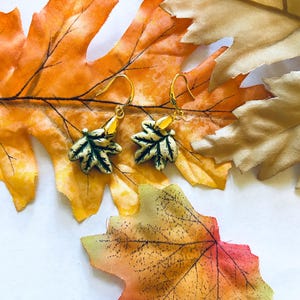 May include: Gold-toned leaf earrings with green and cream accents, displayed on a backdrop of colorful autumn leaves. The earrings feature a detailed leaf design, perfect for fall fashion.