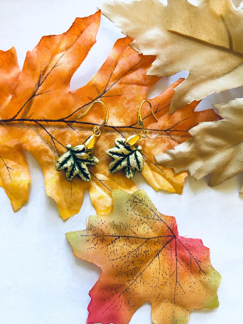 May include: Gold-toned leaf earrings with green and cream accents, displayed on a backdrop of colorful autumn leaves. The earrings feature a detailed leaf design, perfect for fall fashion.
