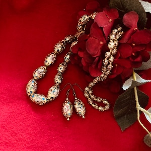 Puede incluir: Un collar y pendientes de oro con cuentas ovaladas decoradas con flores de esmalte de colores. Las joyas se muestran sobre un fondo rojo con un ramo de flores rojas.