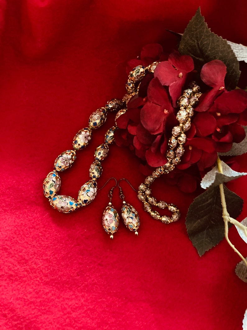 Puede incluir: Un collar y pendientes de oro con cuentas ovaladas decoradas con flores de esmalte de colores. Las joyas se muestran sobre un fondo rojo con un ramo de flores rojas.