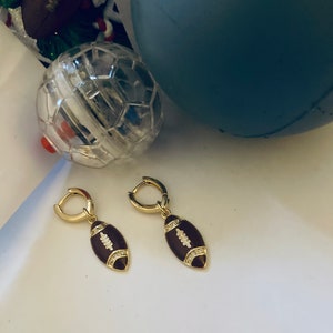 May include: A pair of gold hoop earrings with a brown football charm. The football charm has white accents and small clear stones.