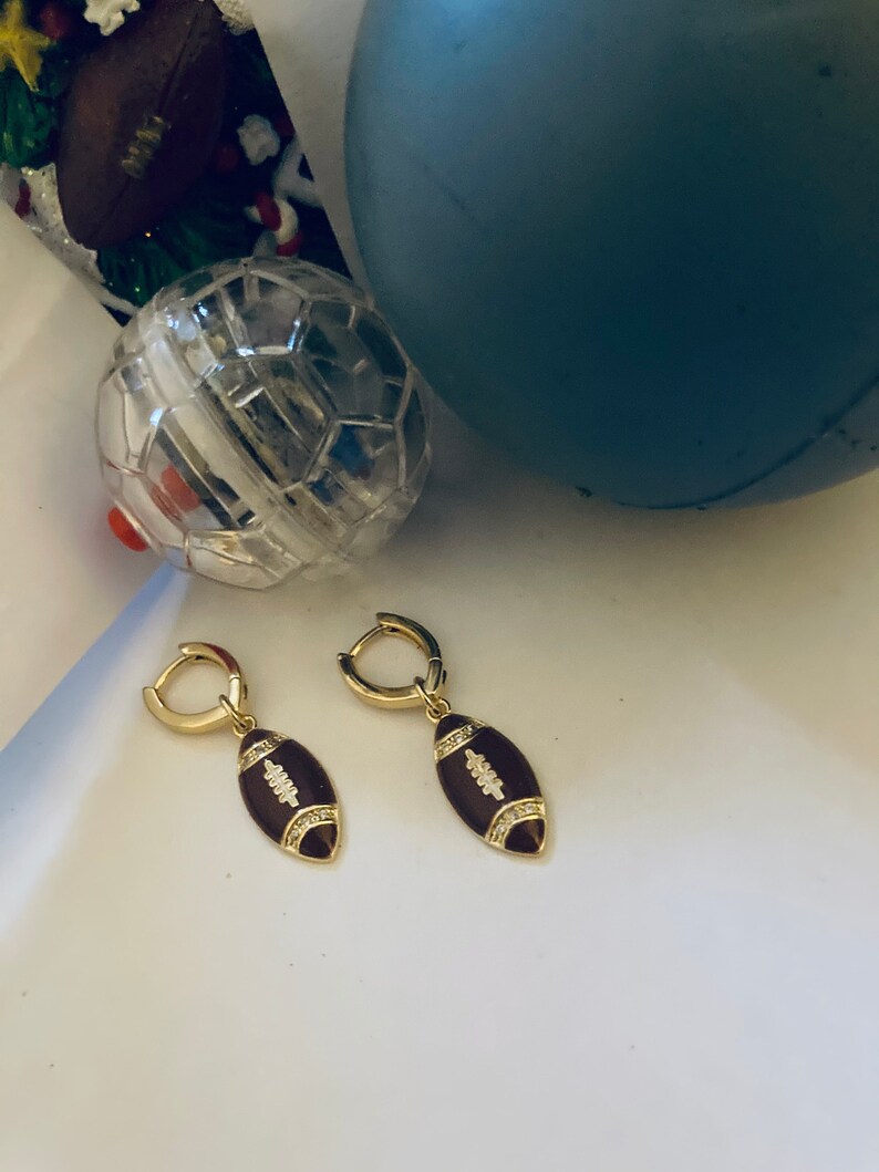 May include: A pair of gold hoop earrings with a brown football charm. The football charm has white accents and small clear stones.