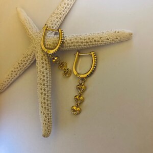 May include: A pair of gold hoop earrings with a dangling chain of three heart charms. The earrings are on a white starfish.