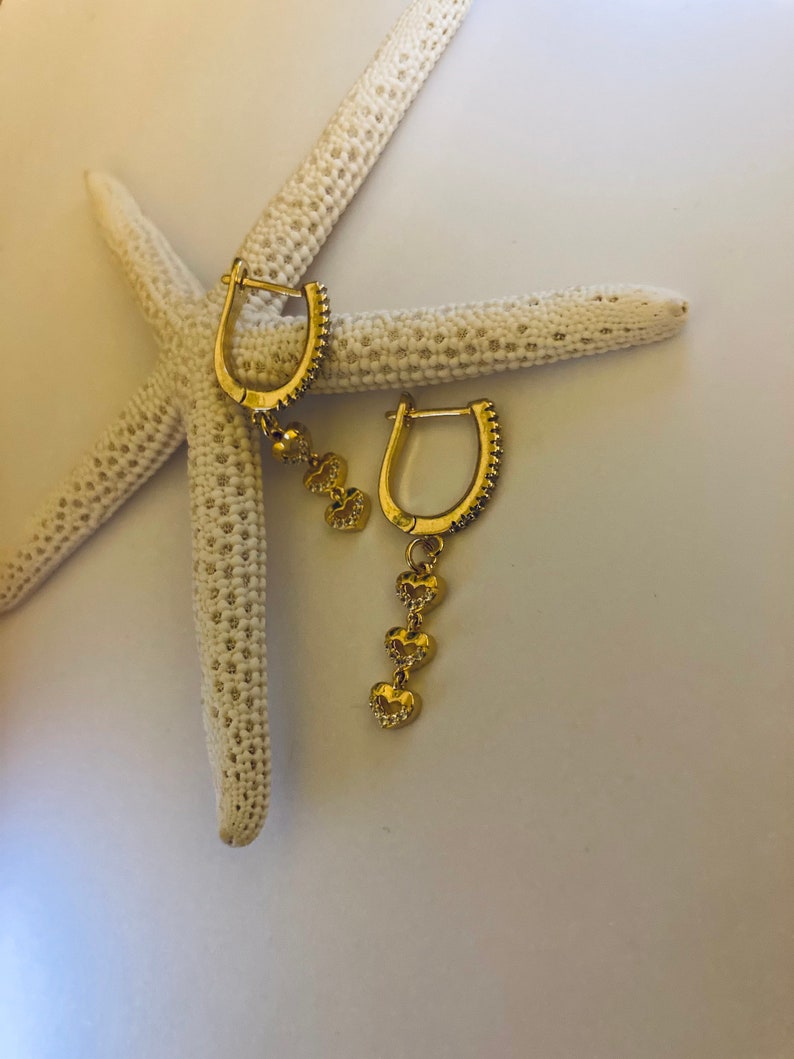 May include: A pair of gold hoop earrings with a dangling chain of three heart charms. The earrings are on a white starfish.