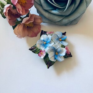 May include: A porcelain brooch featuring a cluster of blue and pink flowers with green leaves. The flowers have a delicate, hand-painted look.