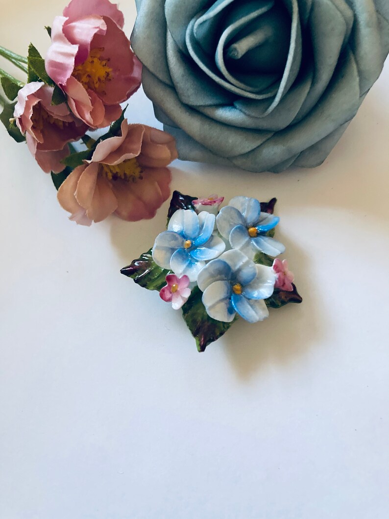 May include: A porcelain brooch featuring a cluster of blue and pink flowers with green leaves. The flowers have a delicate, hand-painted look.