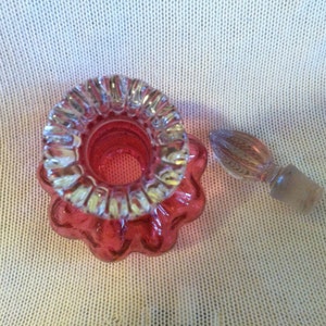 May include: A vintage, ruby red perfume bottle with a clear glass stopper. The bottle has a ruffled rim and a flower-like base. The stopper is clear with a textured design. The bottle is likely made of glass.