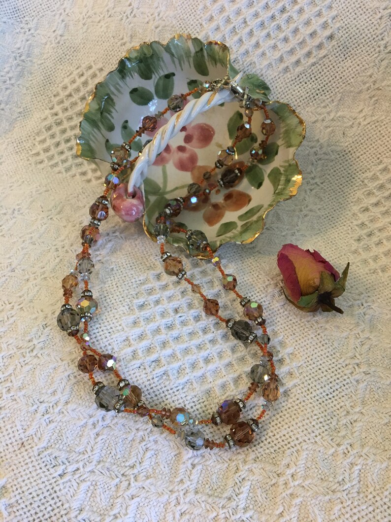 May include: A double strand necklace with iridescent brown and clear glass beads. The necklace is displayed in a white ceramic dish with a floral design.