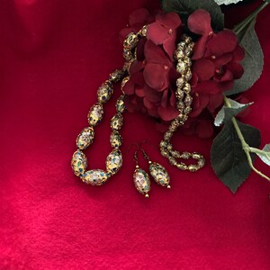 Puede incluir: Un collar y pendientes de oro con dise&ntilde;os florales de esmalte de colores. El collar est&aacute; hecho de cuentas ovaladas y los pendientes son de estilo colgante. Las joyas se muestran sobre un fondo de tela roja con una flor roja.