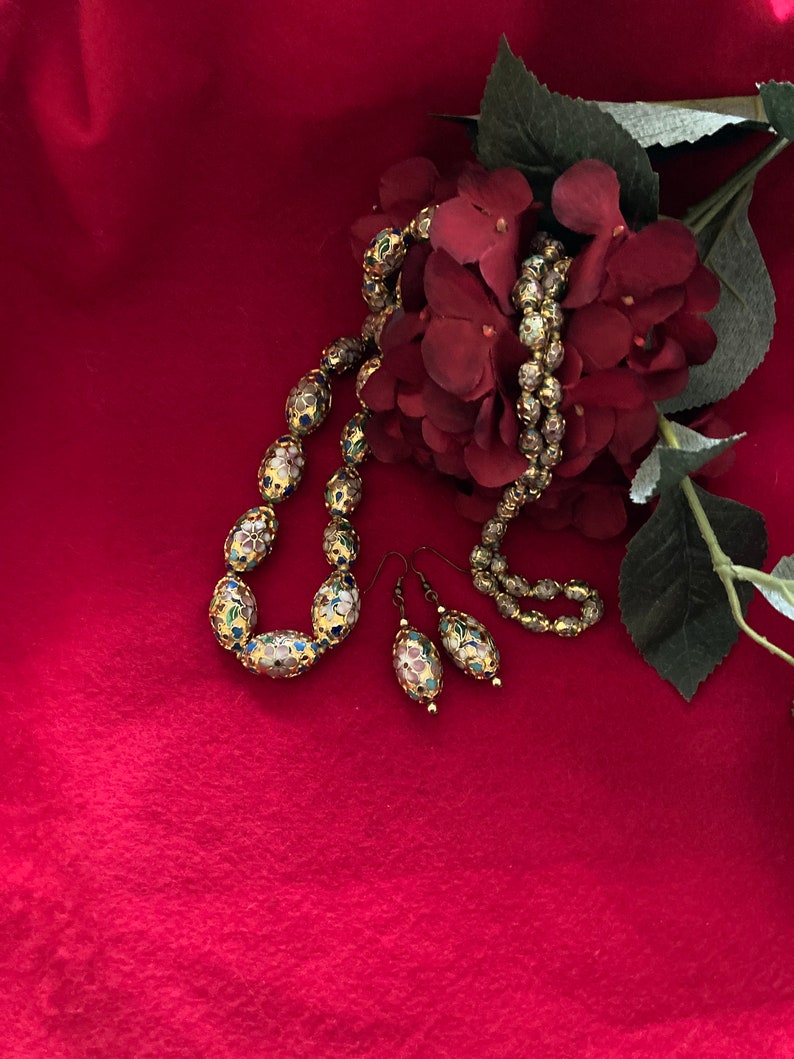 Puede incluir: Un collar y pendientes de oro con dise&ntilde;os florales de esmalte de colores. El collar est&aacute; hecho de cuentas ovaladas y los pendientes son de estilo colgante. Las joyas se muestran sobre un fondo de tela roja con una flor roja.