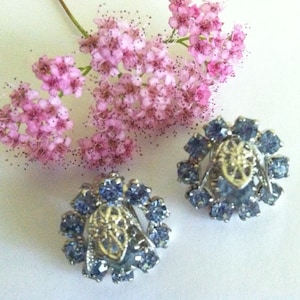 May include: A pair of silver tone earrings with a floral design. The earrings are made of metal and have a cluster of light blue rhinestones in the center. The rhinestones are surrounded by a silver metal frame.