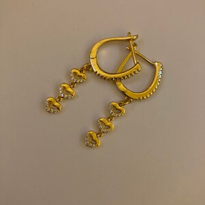 May include: Gold hoop earrings with a dangling chain of five small heart charms, each set with clear cubic zirconia stones.
