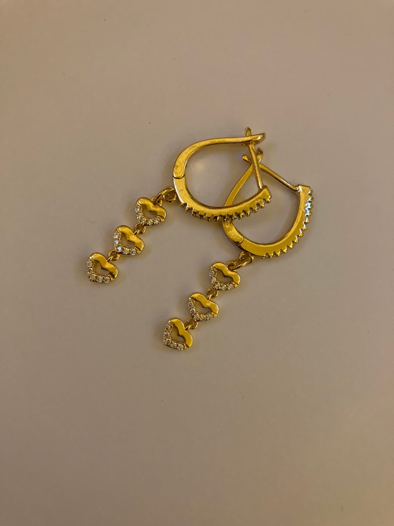 May include: Gold hoop earrings with a dangling chain of five small heart charms, each set with clear cubic zirconia stones.