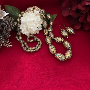 Puede incluir: Un collar y pendientes de oro con cuentas ovaladas decoradas con motivos florales coloridos. Las cuentas son de vidrio y tienen un acabado brillante.