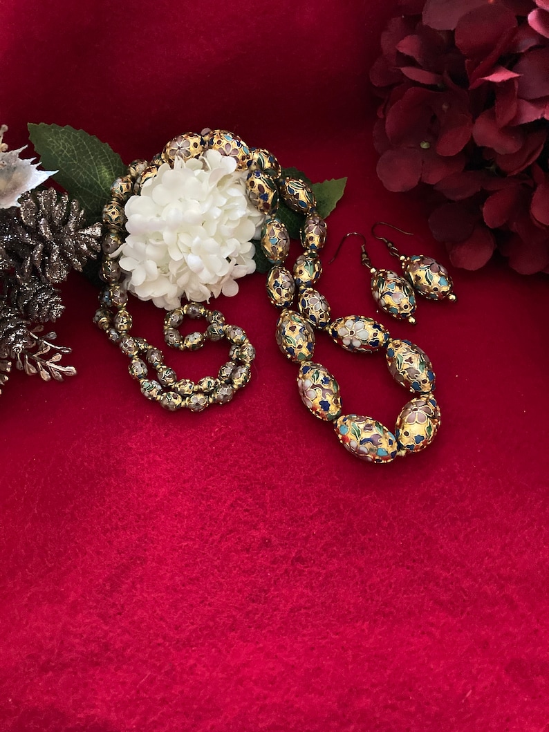 Puede incluir: Un collar y pendientes de oro con cuentas ovaladas decoradas con motivos florales coloridos. Las cuentas son de vidrio y tienen un acabado brillante.